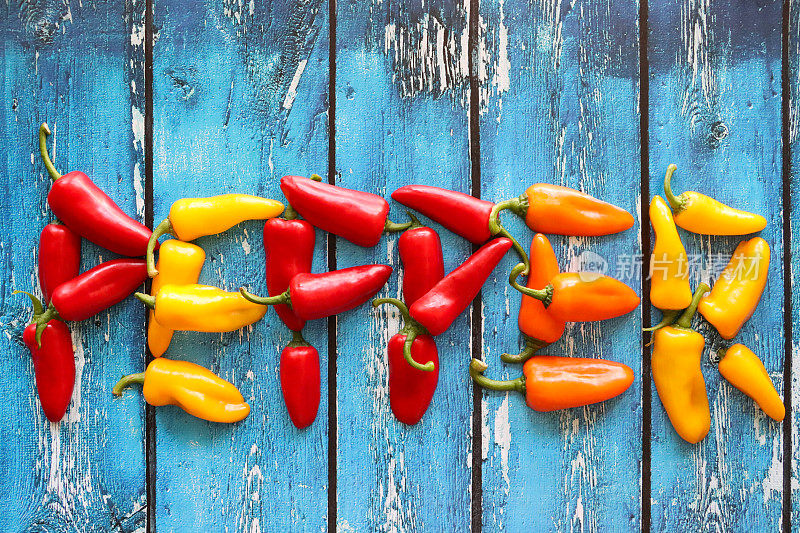 “胡椒”这个词的特写图像是由红色、黄色和橙色的迷你辣椒(Capsicum annuum)对半组成的，蓝色木纹背景，高架视图